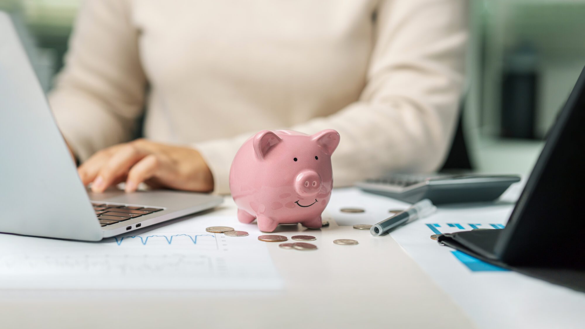Person working at a desk with a laptop, papers, coins, and a pink piggy bank, suggesting financial planning or budgeting.