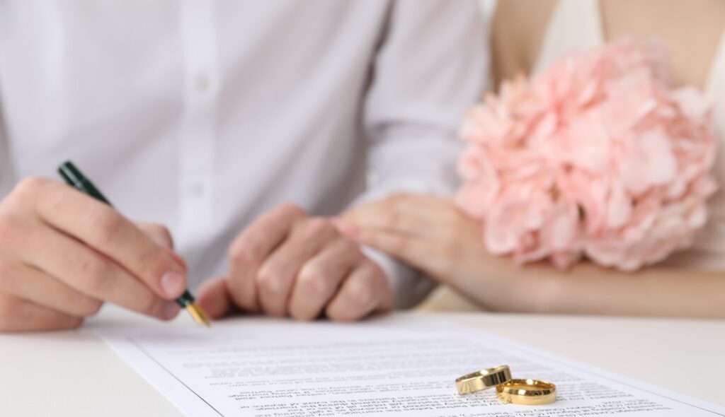 A person signs a marriage contract at a table with two rings and a bouquet of pale pink flowers in the foreground, guided by a prenup attorney in Dayton.