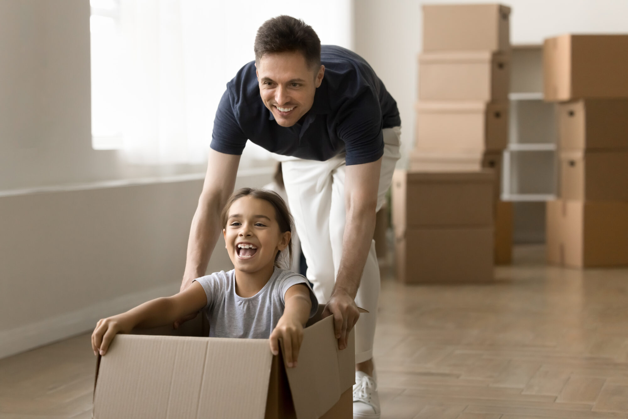 A man pushes a smiling child sitting inside a cardboard box, with moving boxes stacked in the background of a bright room—capturing the joy of new beginnings, perhaps after guidance from a Dayton relocation attorney.